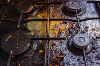 Dört dairesel ocağı olan paslanmış, kirli bir gaz ocağı kapağı. Yüzey, yanmış kahverengi kırıntılar ve sarı kalıntılar da dahil olmak üzere besin kalıntılarıyla kaplıdır.