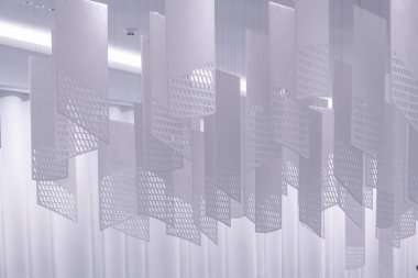 Tavandan sarkan bir dizi geometrik desenli kağıt fener. Fenerler, şebeke benzeri bir dizaynla dikdörtgen şeklindedir. Beyaz, hafif ışıklandırılmış arka plan üzerine karmaşık gölgeler bırakırlar.