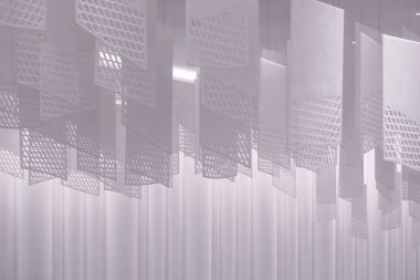 Tavandan sarkan bir dizi geometrik desenli kağıt fener. Fenerler, şebeke benzeri bir dizaynla dikdörtgen şeklindedir. Beyaz, hafif ışıklandırılmış arka plan üzerine karmaşık gölgeler bırakırlar.