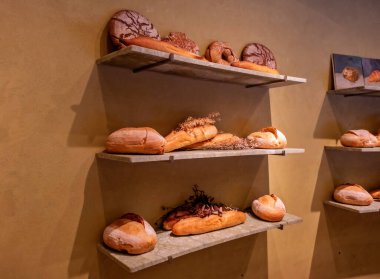 Görüntü, üç rafta çeşitli zanaatkar ekmeklerin bulunduğu şık, minimalist bir fırın görüntüsünü sergiliyor.