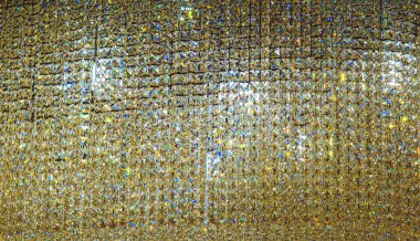 Sayısız küçük, yansıtıcı, altın renkli cam boncuklarla kaplı parıldayan, dokulu bir duvar. Boncuklar yoğun bir şekilde paketlenmiş, ışıltılı, mozaik bir etki yaratıyor.