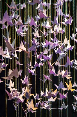 Kağıt origami kuşları siyah arka planda asılı duran iplere bağlı. Kağıt origami kuşları boyut ve oryantasyon olarak farklılık göstererek dinamik, üç boyutlu bir etki yaratırlar.