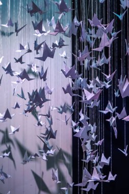 Kağıt origami kuşları siyah arka planda asılı duran iplere bağlı. Kağıt origami kuşları boyut ve oryantasyon olarak farklılık göstererek dinamik, üç boyutlu bir etki yaratırlar.