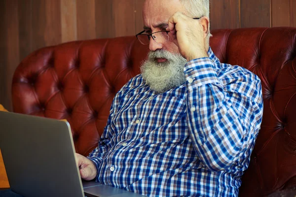 Старик сидит на диване и работает с ноутбуком — стоковое фото