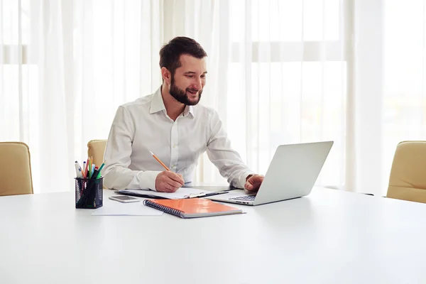 Hombre sonriente trabajando en su portátil y escribiendo algunos datos en un bloc de notas — Foto de Stock