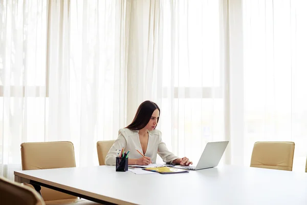 Brunette working on her notebook and writing some data in a pad Royalty Free Stock Photos