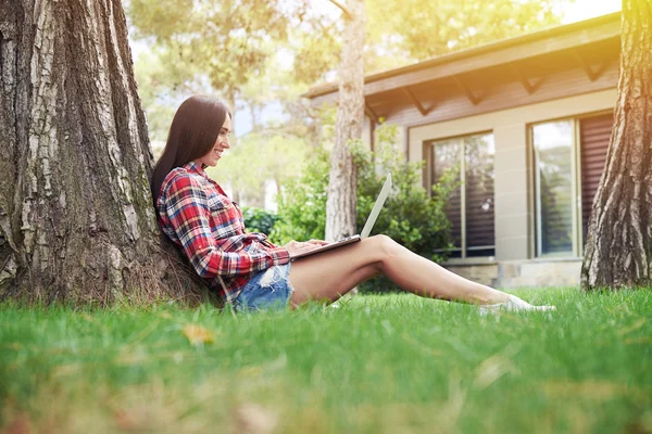 Attraktive junge Frau arbeitet am Laptop unter dem Baum an sonnigen — Stockfoto