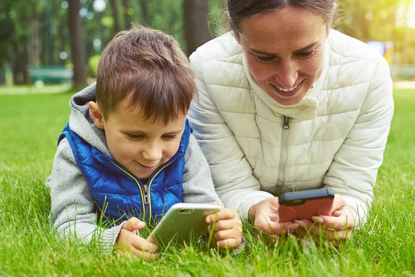Mother and son lying on grass in park and playing on their smar — Stock Photo, Image