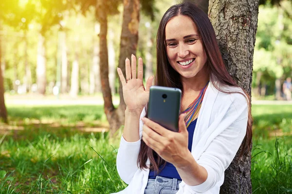 Sorrindo senhora descansando debaixo da árvore e realizando uma chamada de vídeo — Fotografia de Stock