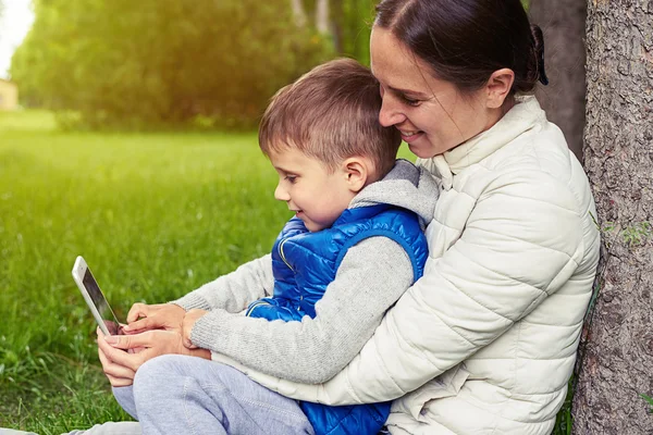 Mujer con hijo viendo algo en el teléfono inteligente bajo el árbol — Foto de Stock