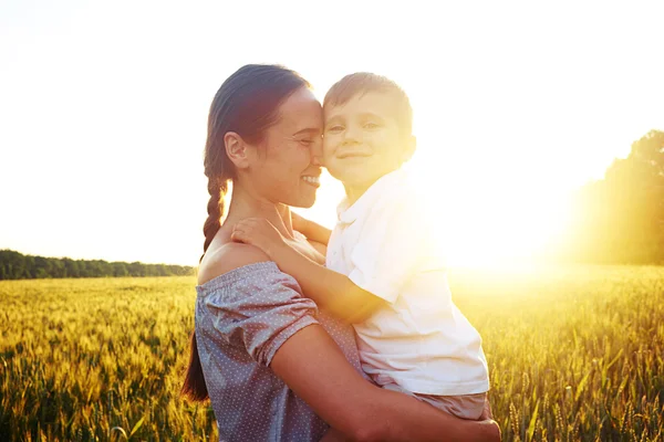 Vacker kvinna som håller liten pojke mot solnedgången i fältet — Stockfoto