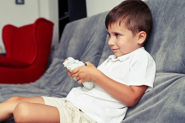 Menino pequeno é arrebatado em jogar um videogame sentado no sofá cinza — Fotografia de Stock