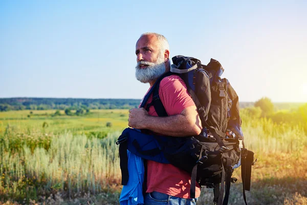 Handsome aged man standing in front of sunlit field carring a ru — Stock Photo, Image