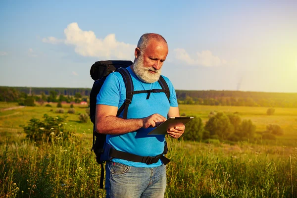 Sporty aged man with rucksack using data tablet against field ba — Stock Photo, Image