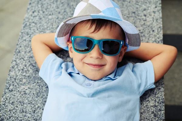 Gros plan du petit garçon dans des lunettes de soleil couché avec les mains sous son h — Photo