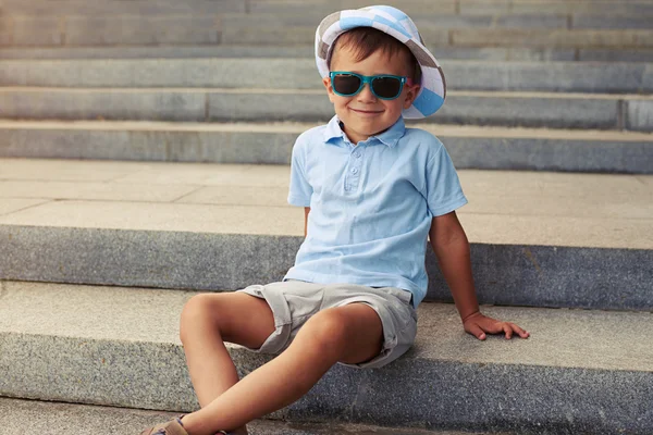 Petit garçon aux lunettes de soleil bleues assis sur des escaliers en béton à l'extérieur — Photo