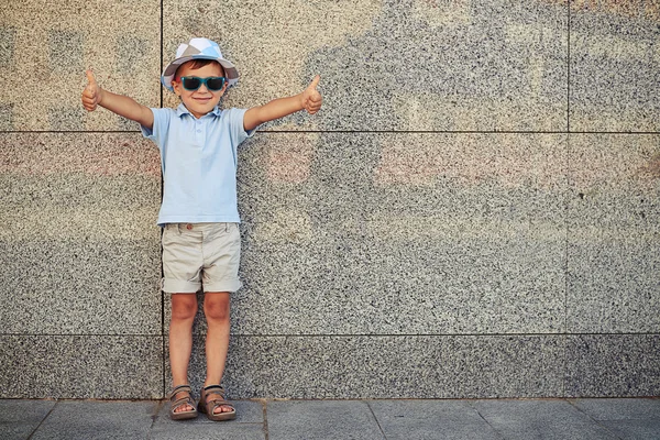 Petit garçon en lunettes de soleil debout près du mur dans la rue et — Photo