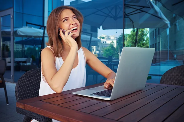 Young woman in street cafe using her laptop and talking on mobi — Stock Photo, Image