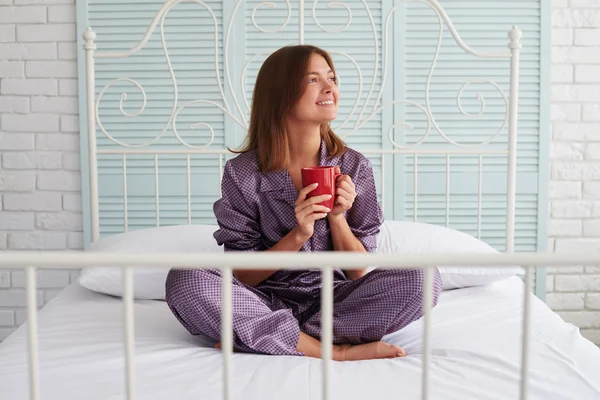 Atrakcyjna dziewczyna w piżamie z filiżanką kawy siedzącej na łóżku a — Zdjęcie stockowe