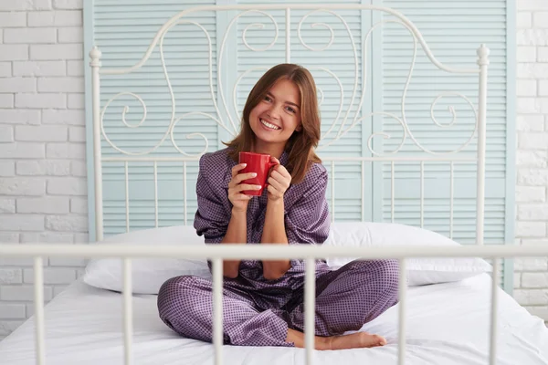 Красивая девушка в пижаме сидит на кровати с красной чашкой и улыбкой — стоковое фото