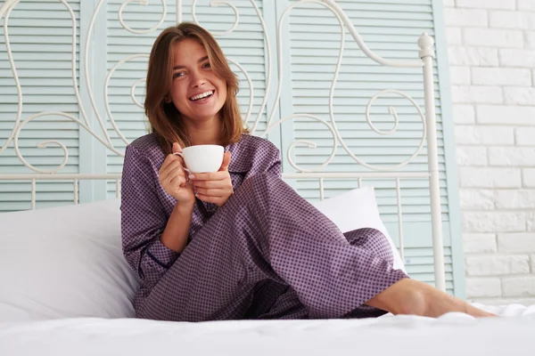 Vacker flicka i natt-kostym sitter vid sängen med en kopp o — Stockfoto