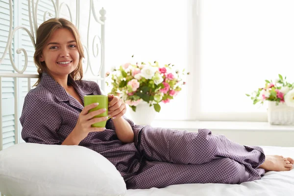 Porträtt av leende vacker kvinna i pyjamas på sängen med en kopp — Stockfoto