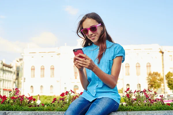 Hermosa adolescente en gafas de sol de color rosa sentado en el lecho de flores de un —  Fotos de Stock