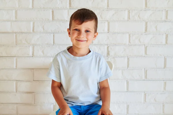 Portrait de garçon mignon souriant assis sur une chaise contre bric blanc — Photo