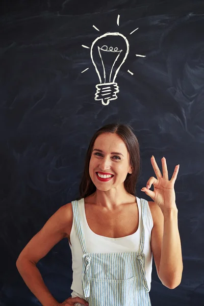 Beautiful female showing ok sign against chalk drawing of light — Stock Photo, Image