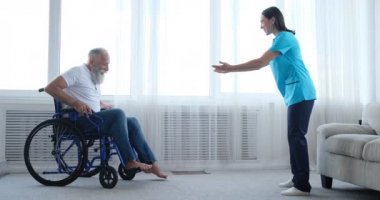 Kadın hemşire engelli yaşlı hastayı tekerlekli sandalyeden kalkmaya teşvik ediyor.