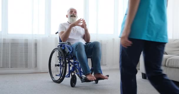 Homem idoso deficiente recusando enfermeira feminina para ajuda e empurrando-se em cadeira de rodas — Vídeo de Stock