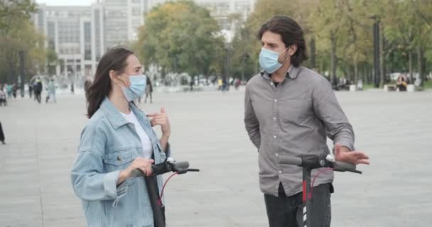 Pareja que usa mascarilla protectora y habla apoyada en scooters electrónicos — Vídeos de Stock