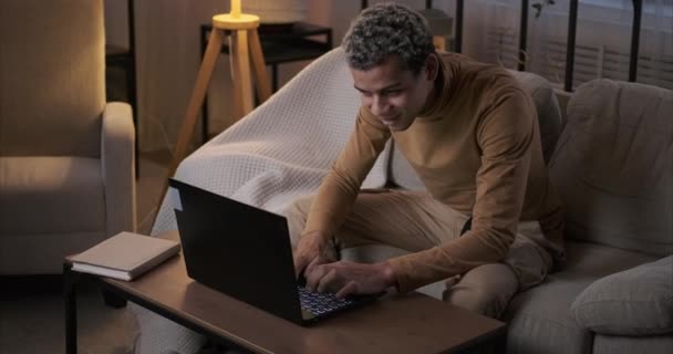 Homem usando laptop sentado no sofá à noite — Vídeo de Stock