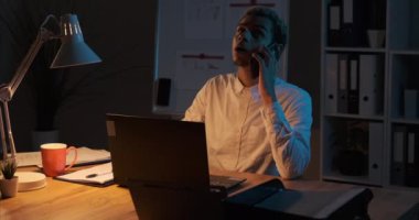 İşadamı bilgisayarını kullanırken belgeleri analiz ediyor ve gece ofisinde cep telefonuyla konuşuyor.