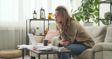 Evrak işleriyle uğraşırken kahve içen ve evde laptop kullanan bir kadın.