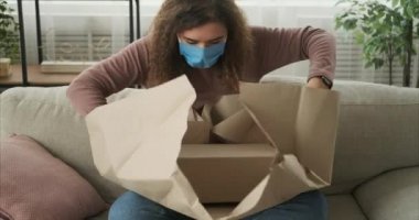 Evde yüz maskesi takan ve paketi açan bir kadın var.