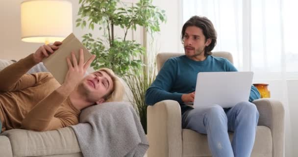 Gay hombre leyendo libro mientras novio es usando laptop en casa — Vídeo de stock