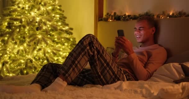 Man liggend op bed met behulp van mobiele telefoon naast kerstboom — Stockvideo