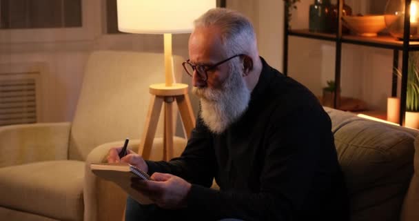 Старший мужчина пишет в книге поздно ночью — стоковое видео