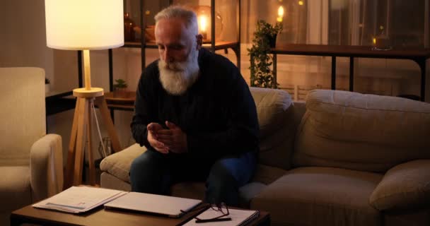 Hombre mayor sentado en el sofá y el uso de la computadora portátil tarde por la noche — Vídeos de Stock