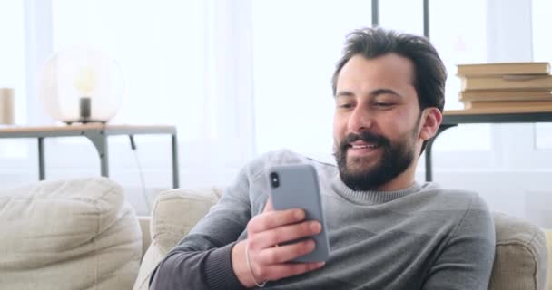 Szczęśliwy młody człowiek za pomocą telefonu komórkowego podczas relaksu na kanapie — Wideo stockowe
