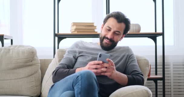 Hombre joven usando el teléfono móvil mientras se relaja en el sofá — Vídeo de stock