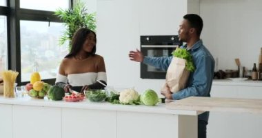 Adam alışveriş torbasında yiyecek getiriyor, karısı da mutfakta yemek hazırlıyor.