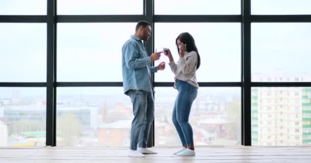 Boldog pár, akik borospoharat tartanak és táncolnak az ablak előtt. — Stock videók
