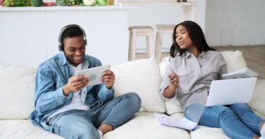 Dizüstü bilgisayarda çalışan kadının dikkati, kulaklıklı kocası tarafından dijital tablette oyun oynarken dağılıyor.