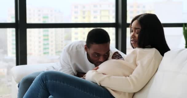 Hombre acariciando y besando vientre de su esposa embarazada sentado en sofá — Vídeos de Stock
