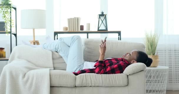 Mujer feliz usando el teléfono móvil y acostado en el sofá — Vídeo de stock