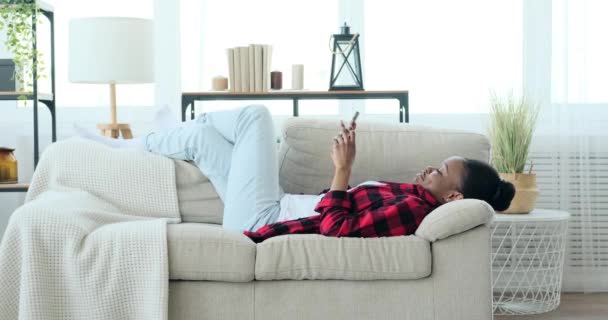 Happy Afro-Amerikaanse vrouw liggend op de bank en met behulp van mobiele telefoon — Stockvideo