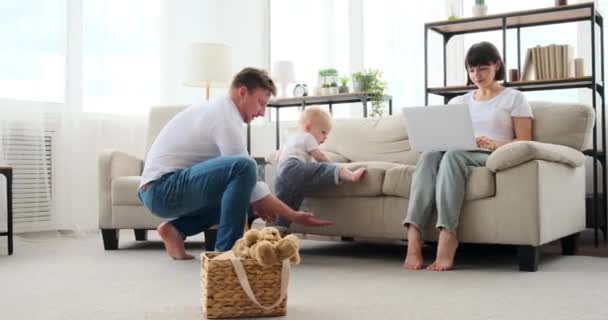 Bambino figlio cercando di salire sul divano con l'aiuto del padre mentre la madre sta lavorando sul computer portatile — Video Stock
