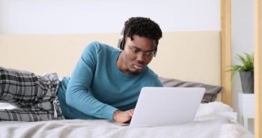 Genç adam dizüstü bilgisayar kullanıyor ve kulaklıkla yatakta müzik dinliyor. 